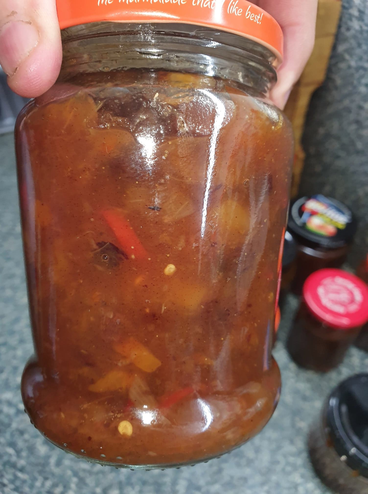 A hand holding a jar of finished chutney, showing mango pieces and strips of red chilli